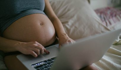 Closeup Of A Young Pregnant Woman Working On Laptop Sitting On Bed.